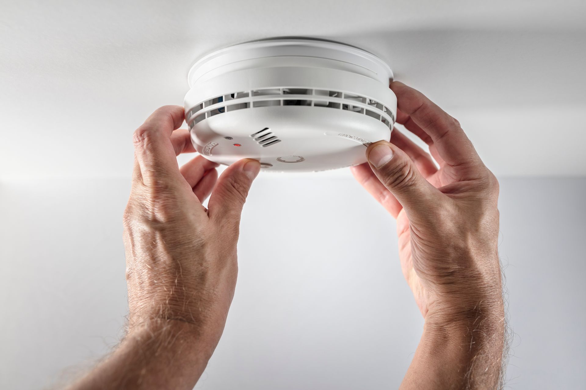 Hands holding a white smoke detector against a light background.