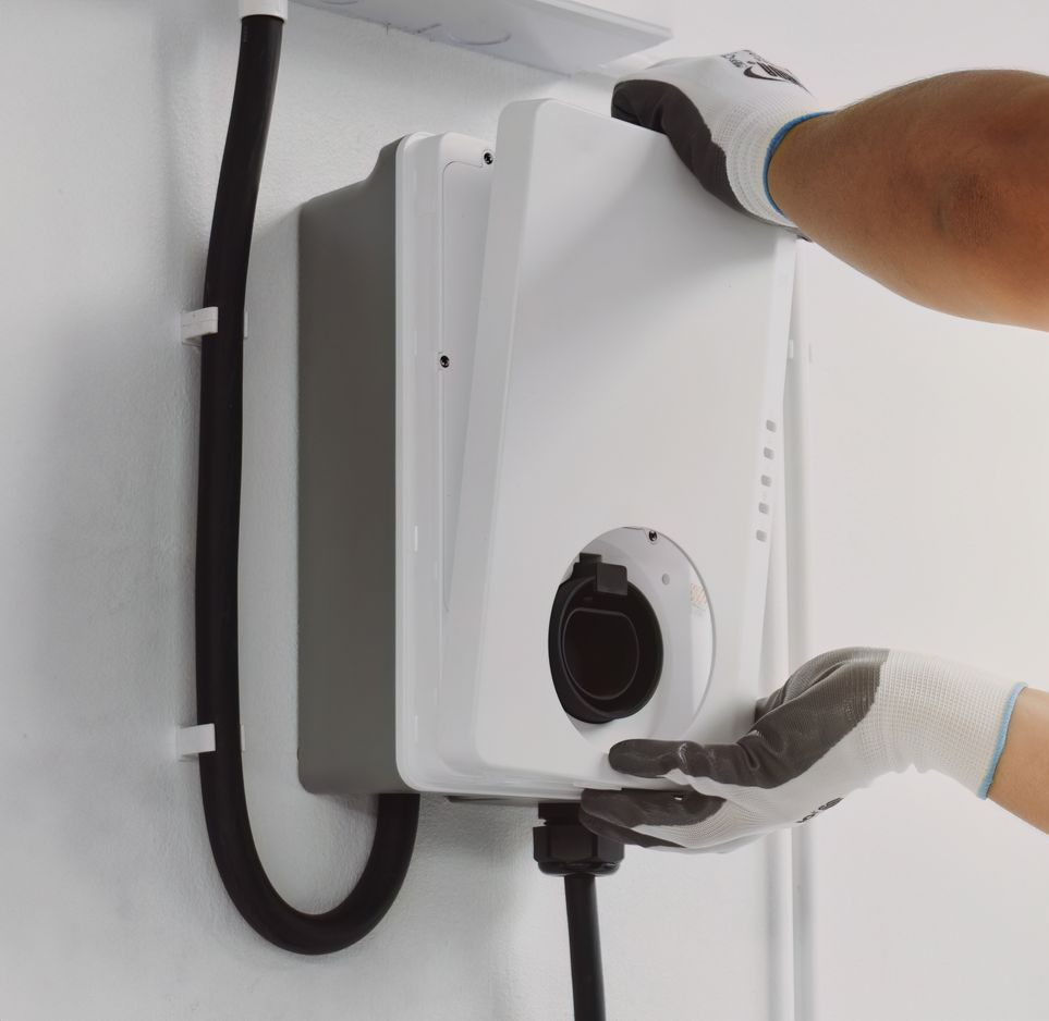 Gloved hands installing a white wall-mounted electric vehicle charger with black cable.