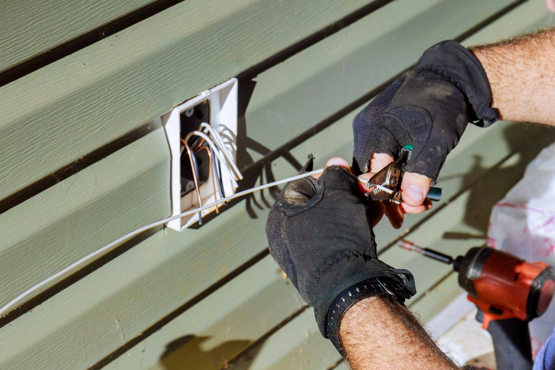 Gloved hands wiring a wall outlet on green siding, with a drill nearby