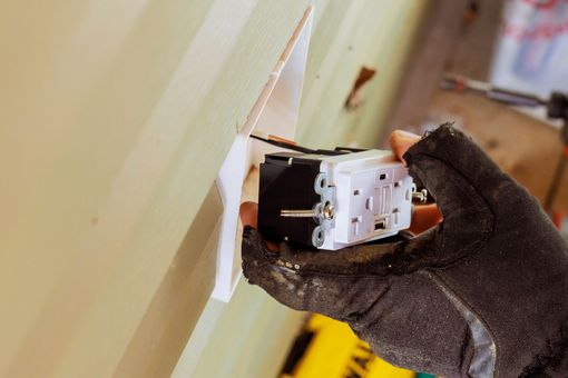 Gloved hand installing a white electrical outlet into a wall opening