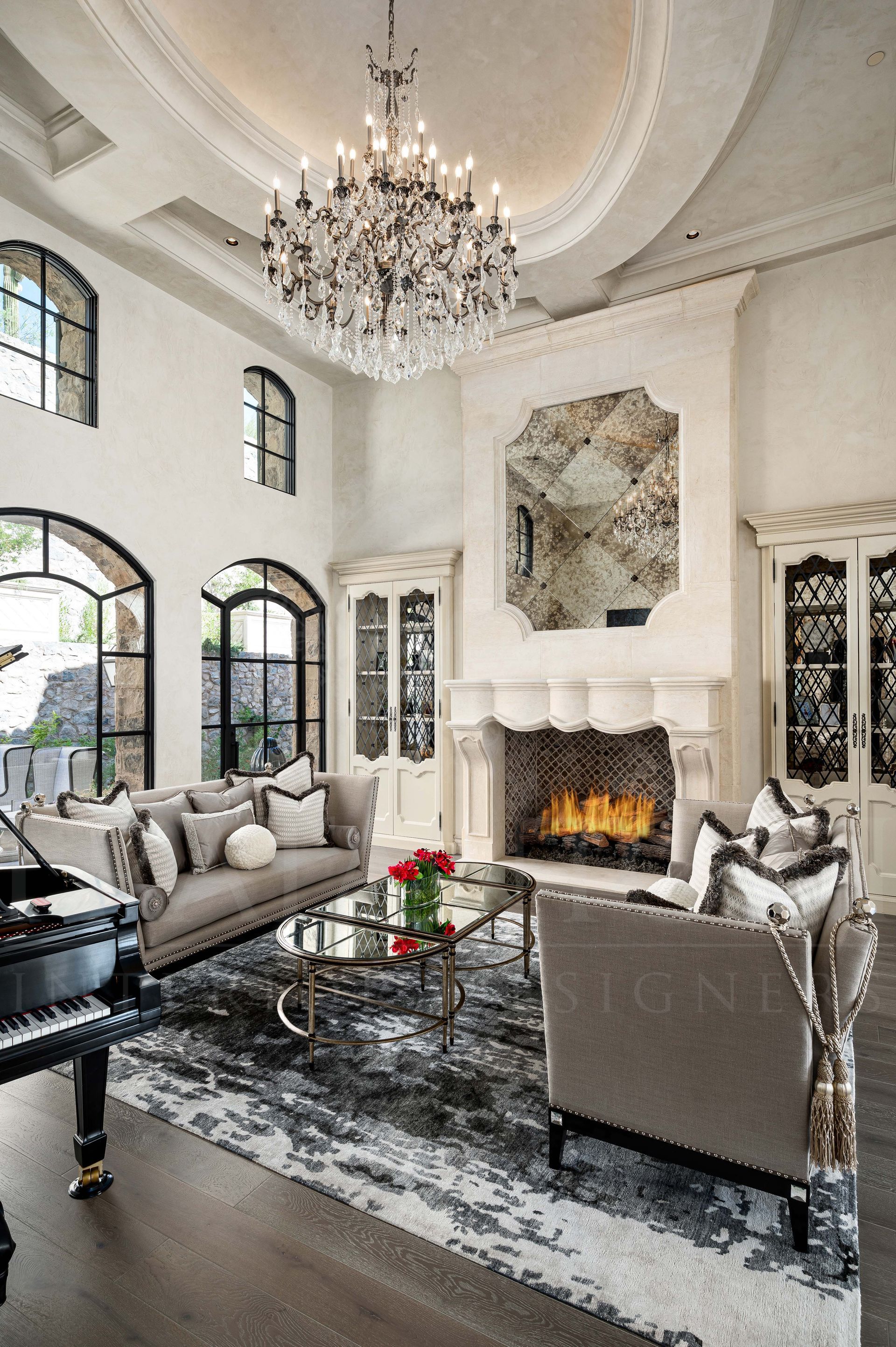French luxury grand living room featuring a crystal chandelier, fireplace, arched windows, and grey furniture on a patterned rug.