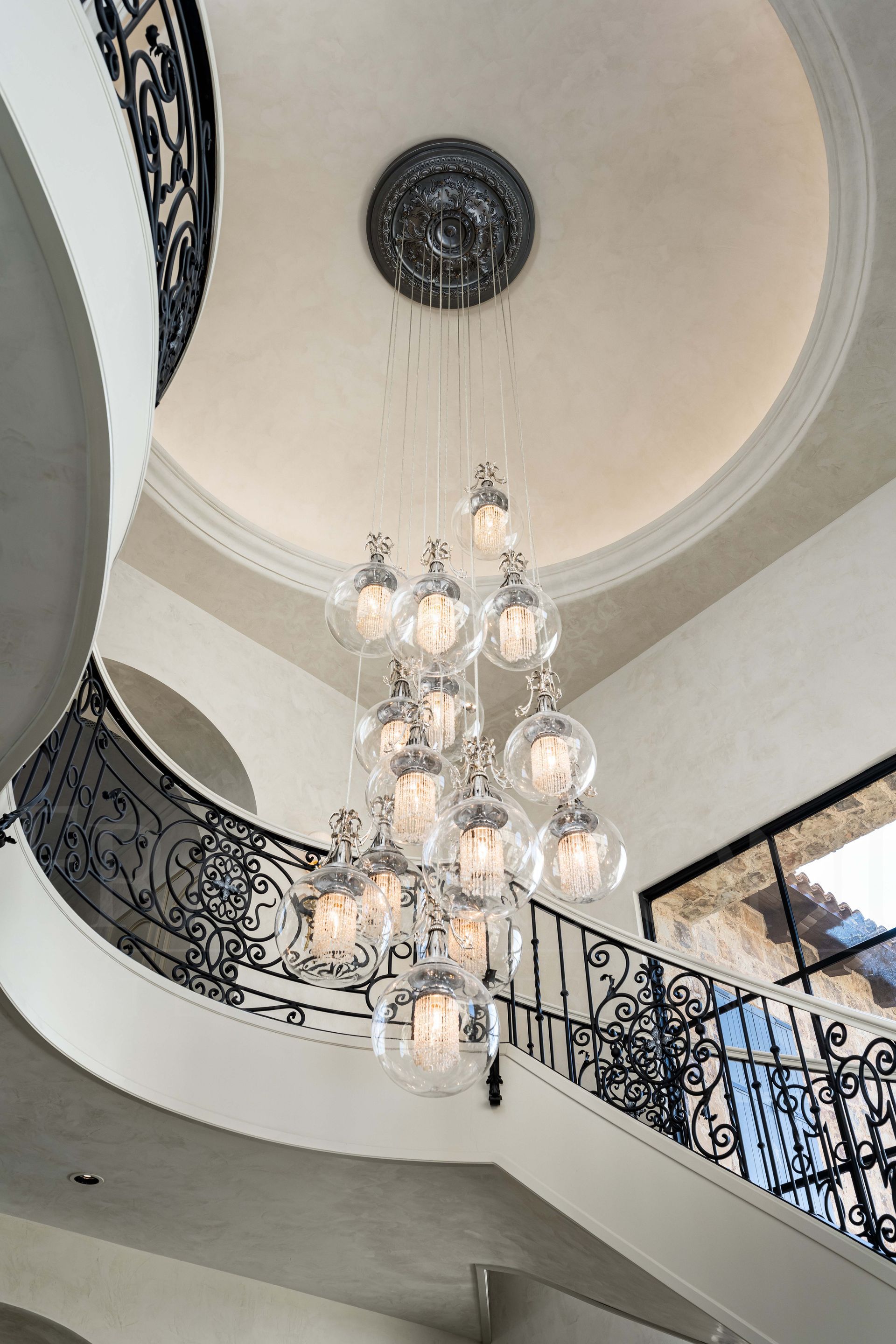 A custom tiered, multi-globe crystal chandelier hangs in the center of a grand staircase with ornate wrought-iron railings.