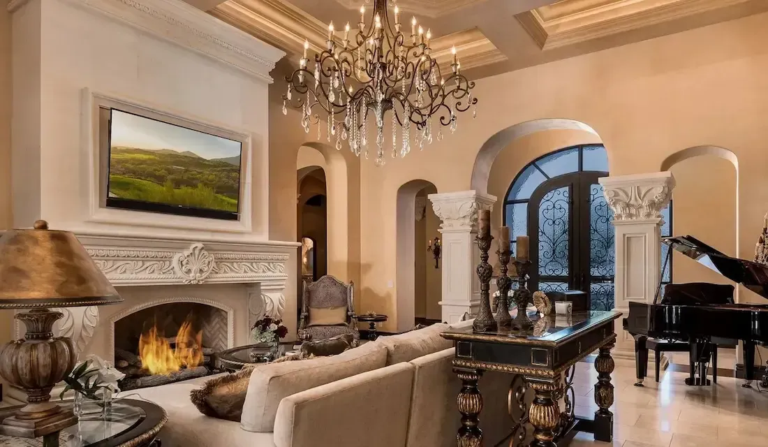 Elegant living room with fireplace, chandelier, piano, and arched doorways, beige and gold tones.