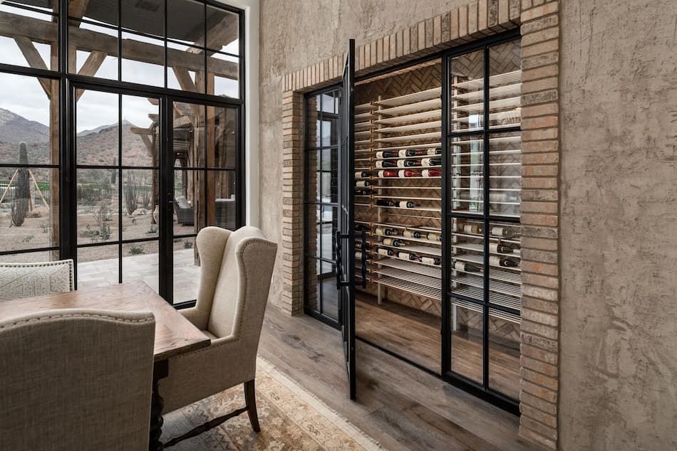 Dining room with large windows, brick wine cellar behind glass doors, and upholstered chairs.
