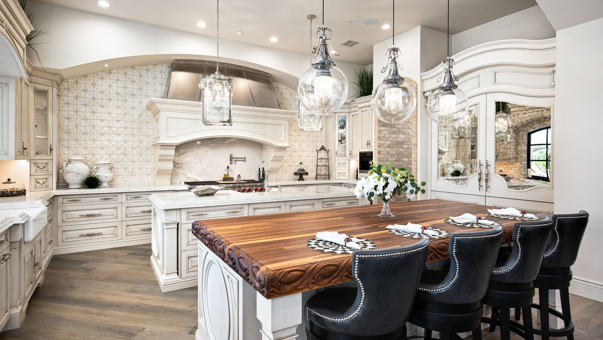A grand, white-toned French chateau kitchen with a large wooden island, ornate cabinetry, and hanging glass pendants.