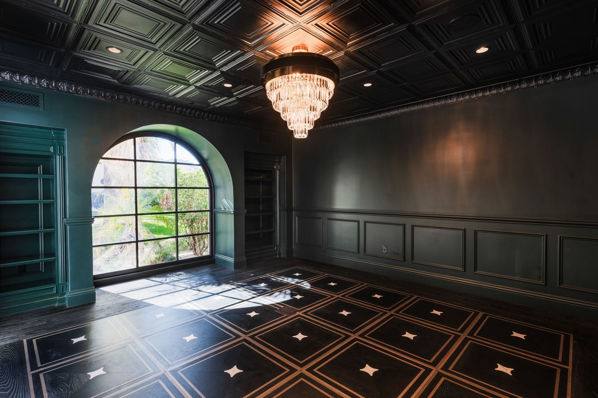 A dark, moody, opulent room with a black ornate ceiling, a crystal chandelier, an arched window, and patterned tile flooring.