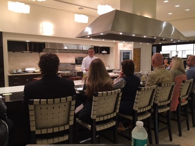 A group of people are sitting in a kitchen watching a man cook