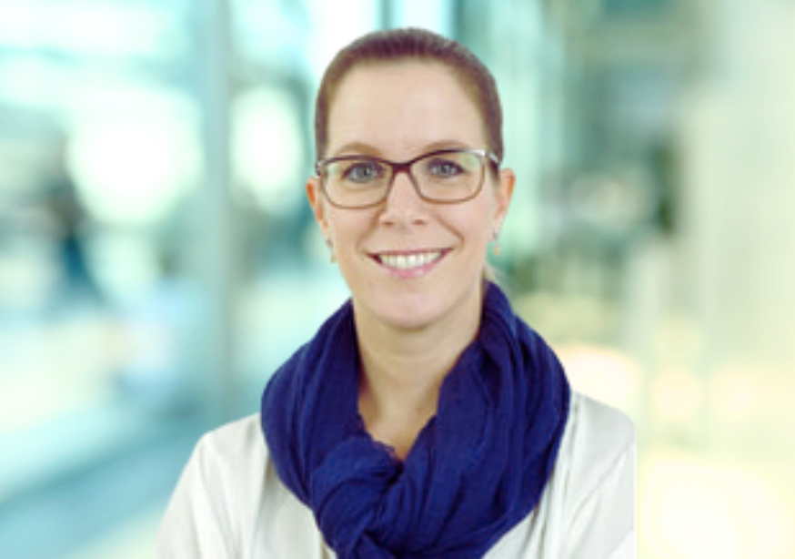 Frau mit Brille und blauem Schal lächelt vor einem verschwommenen Hintergrund.