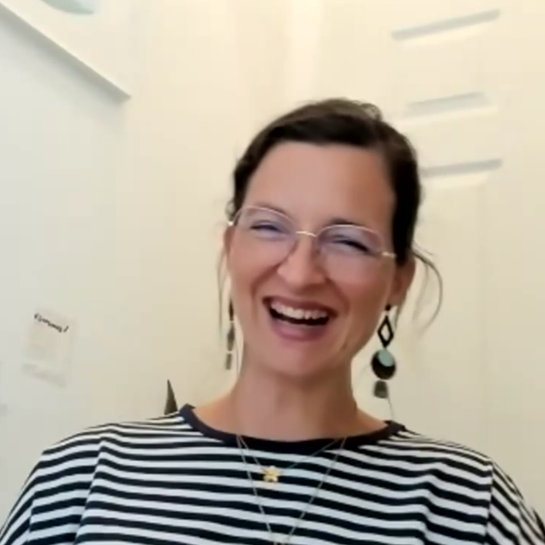 Eine Frau mit Brille, die ein gestreiftes Hemd und baumelnde Ohrringe trägt, lacht in einem hell erleuchteten Raum.