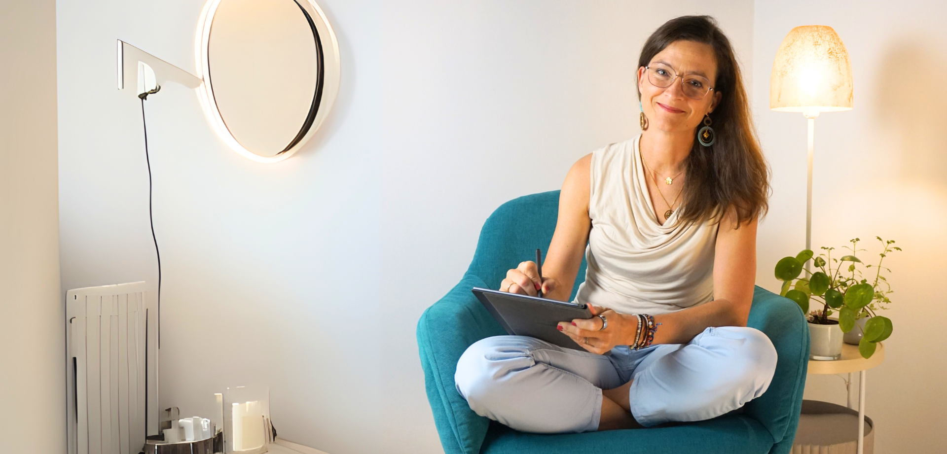 Frau sitzt in blaun Stuhl, hält ein Tablet und lächelt. Sie ist in einem gut beleuchteten Raum mit Lampe & Wanddekoration.