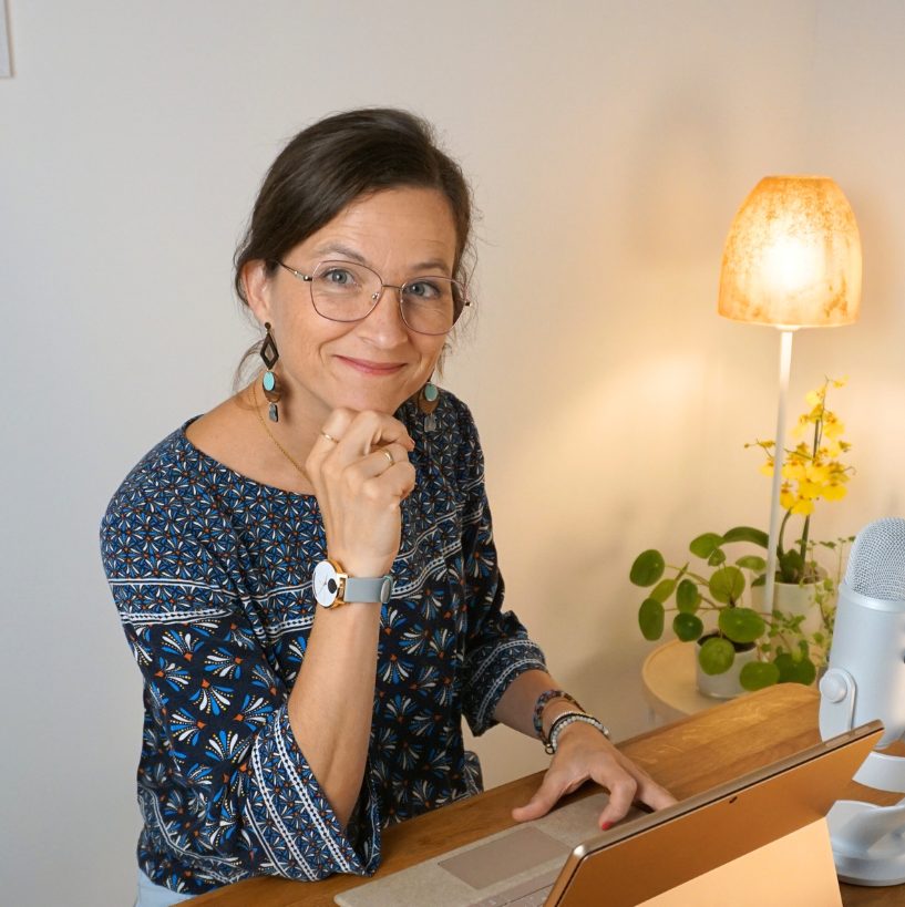 Frau mit Brille lächelt in die Kamera, Hand am Kinn, an einem Schreibtisch mit Laptop und Mikrofon.