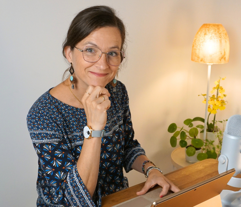 Frau mit Brille lächelt, stützt das Kinn auf die Hand und sitzt mit einem Schreibtisch und einem Mikrofon da.