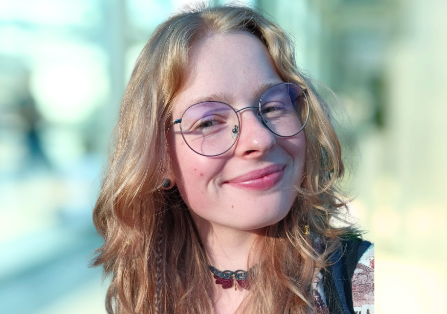 Frau mit lockigem blonden Haar und Brille lächelt. Draußen, unscharfer Hintergrund.