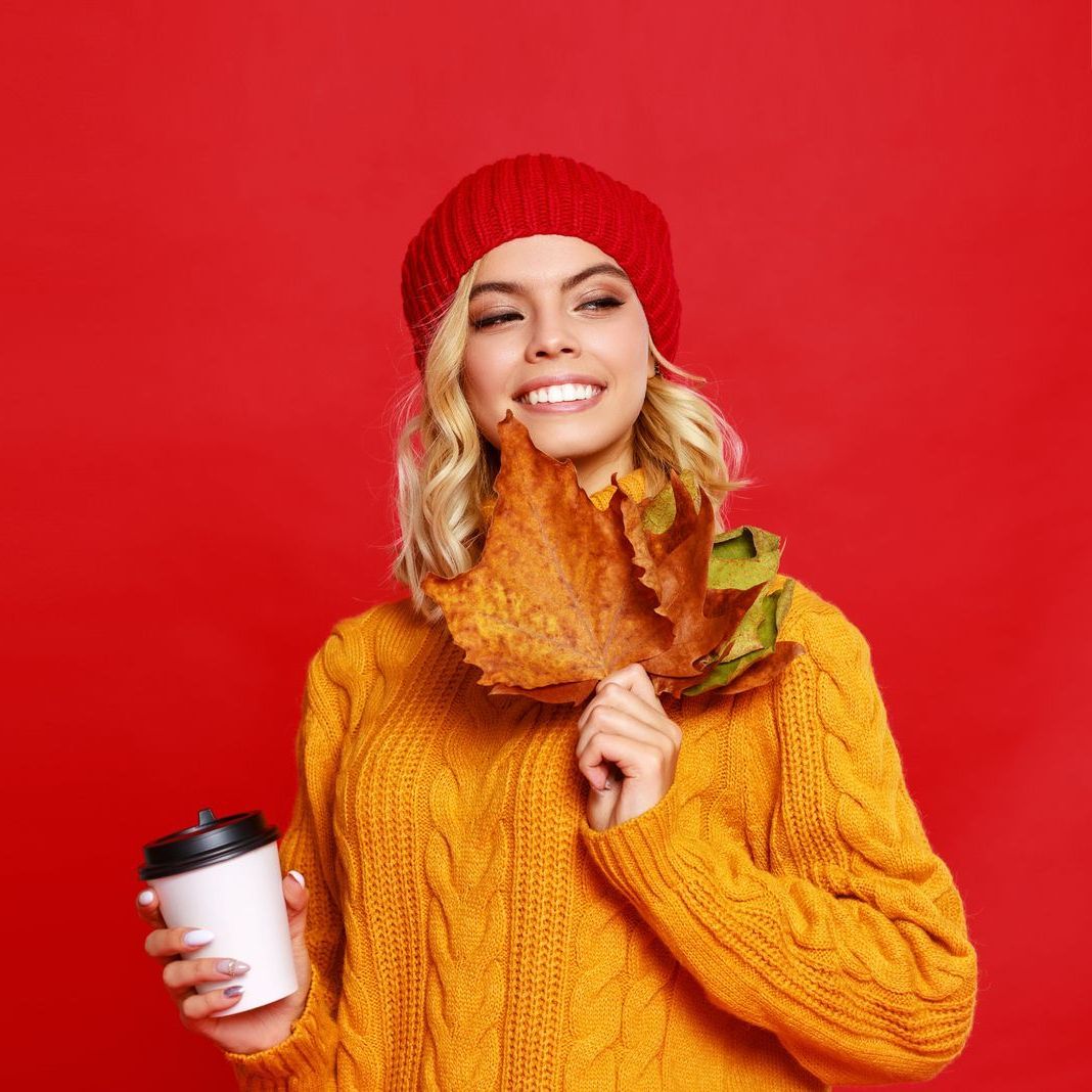 A woman in a yellow sweater and red hat is holding a cup of coffee and a leaf.