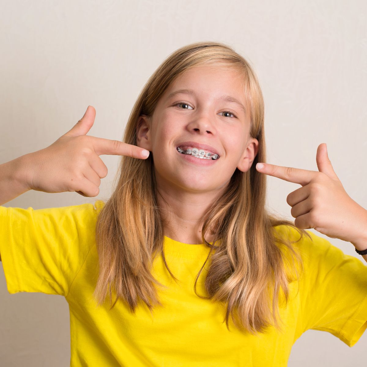 A kid with long hair and a yellow shirt, pointing to their teeth with braces