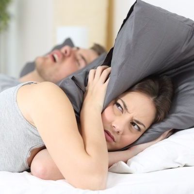 Man snoring in bed, his wife is covering her ears with her pillow