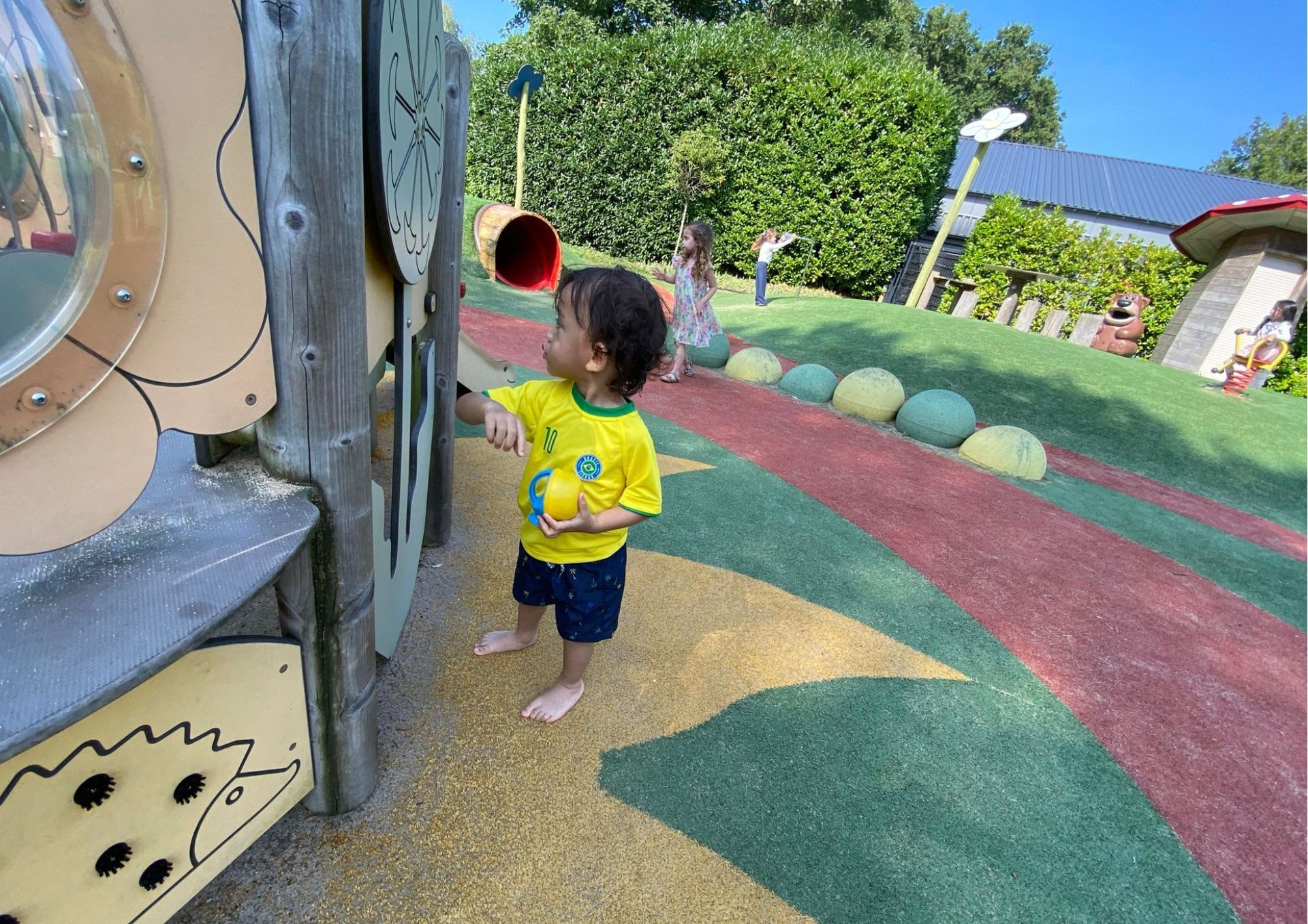 Een jongetje staat op een speelplaats met een gele bal in zijn handen.