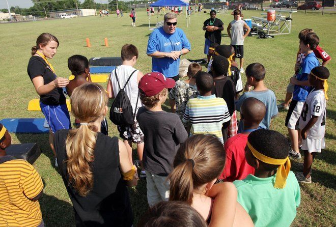 Kids listen to coaches on a grassy field, sunny day.