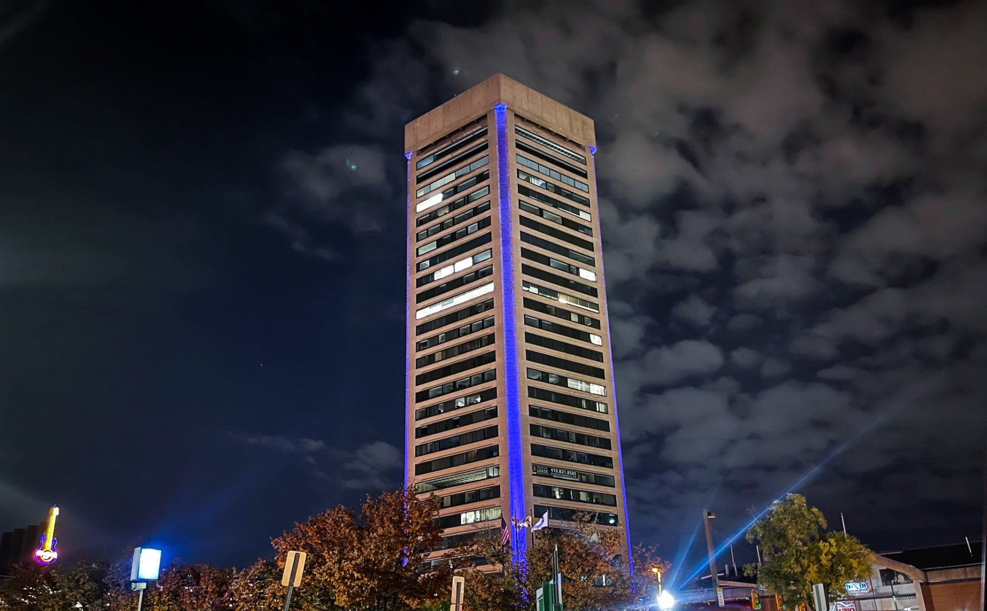 Tall skyscraper illuminated at night with blue accents, trees in the foreground.