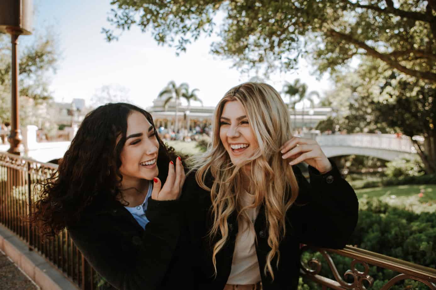 Two smiling women leaning on a railing outdoors; one with dark hair, the other blonde, bridge and trees in background.