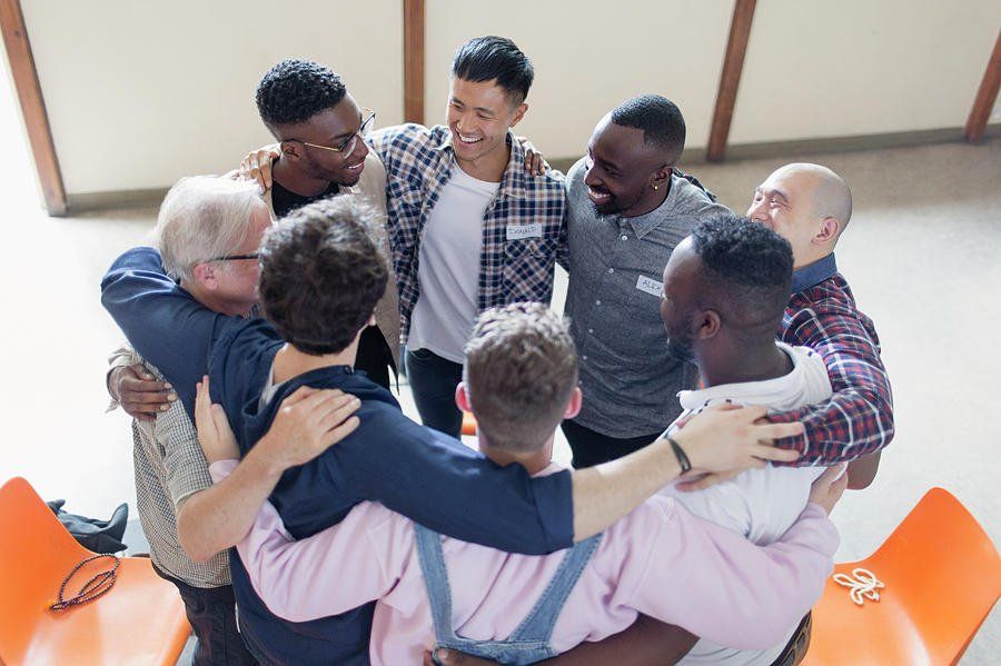 Group of diverse men in a circle, hugging and smiling indoors.