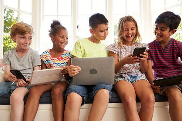 Five diverse kids smiling while using tablets, laptop, and phones by a bright window.