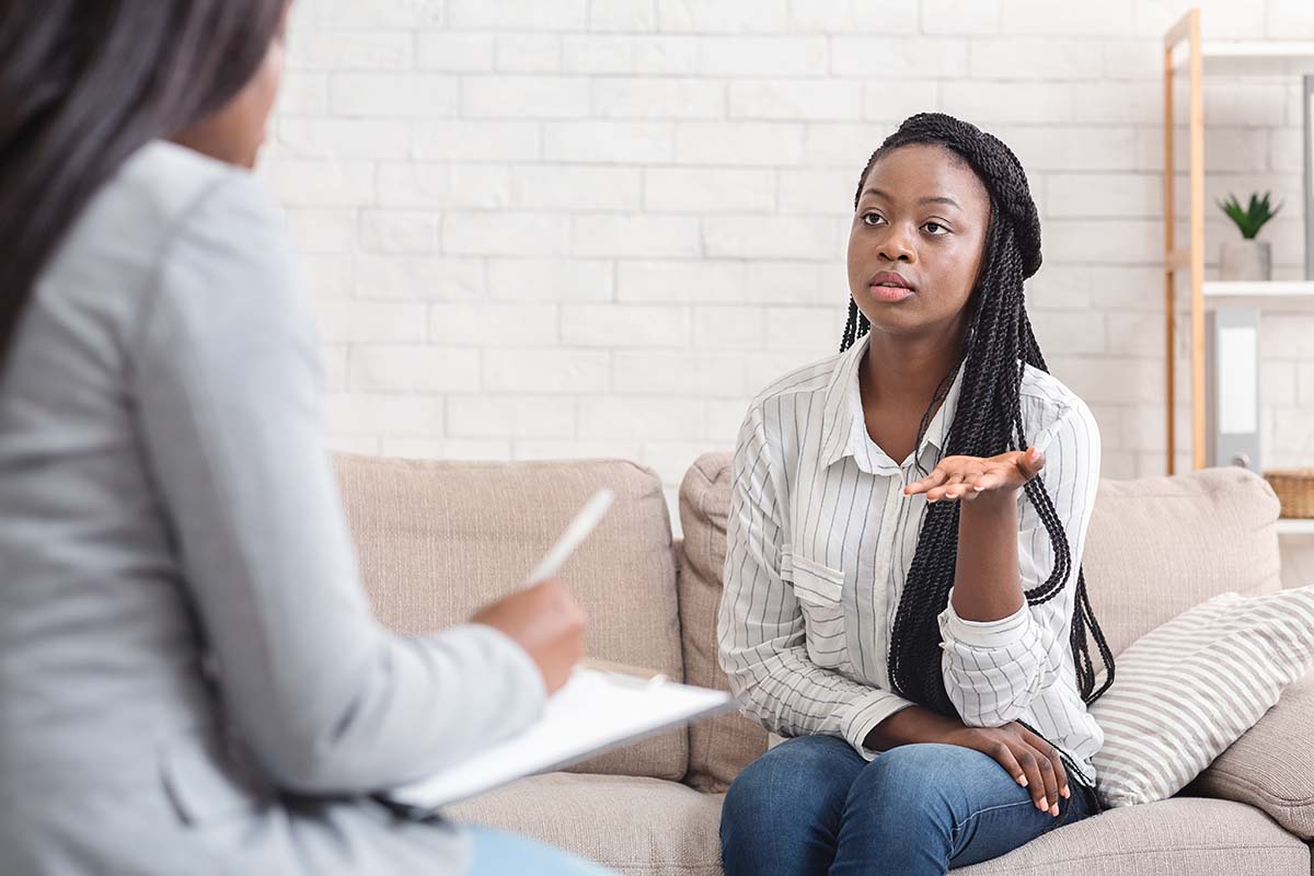Woman with braids talking to a therapist on a couch; therapist takes notes.