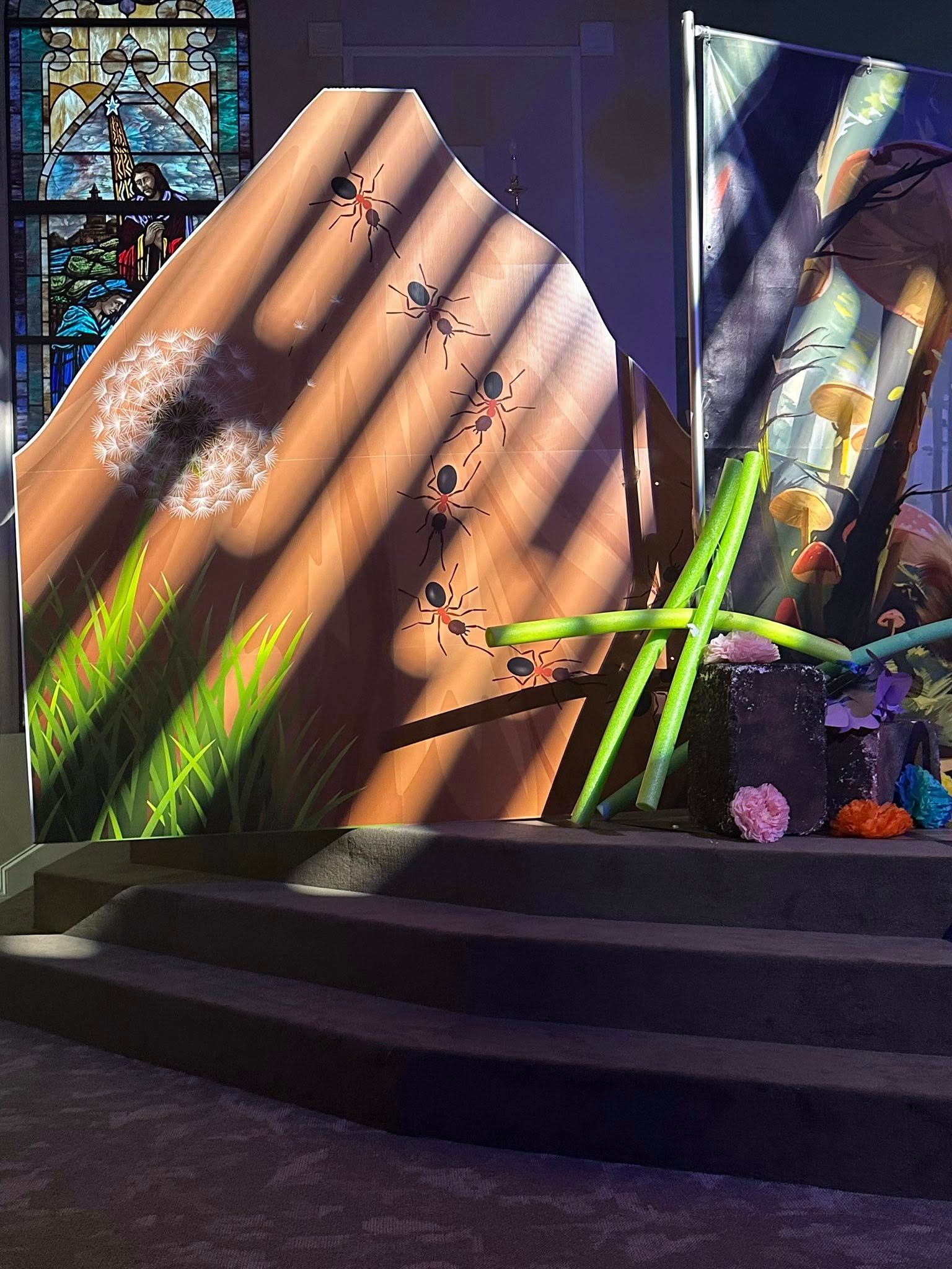 Stage set with wooden panel, painted dandelions, and colorful decorations, including a stained-glass window.