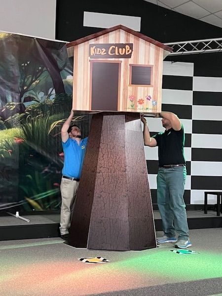 Two men installing a Kid's Club playhouse atop a faux tree trunk. Playhouse is brown, with flowers.