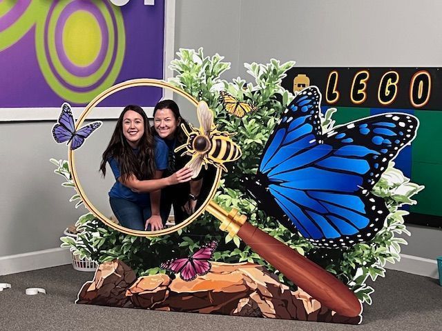 Two women smiling, posing in a butterfly-themed cutout with a magnifying glass.