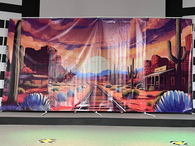 Western-themed backdrop with cacti, mountains, train tracks, and a saloon at sunset.