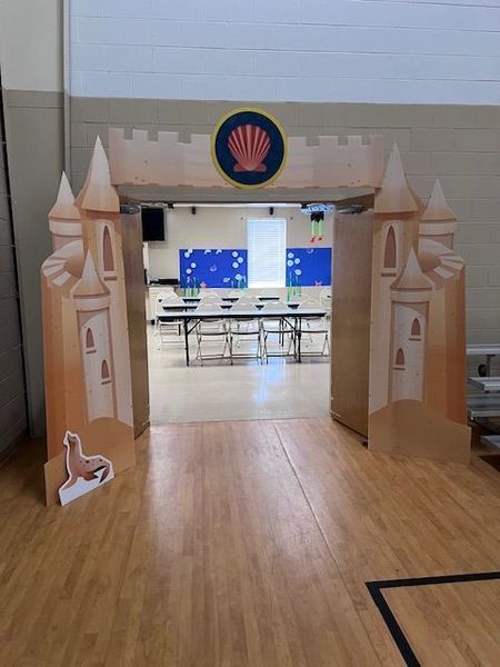 Cardboard castle archway leading into a room with tables and chairs; a seashell emblem is above the opening.