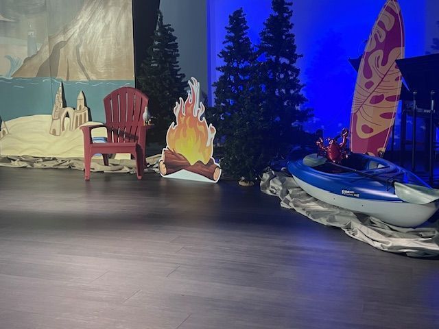 Stage set: Red chair, bonfire cutout, trees, kayak, surfboard, backdrop with sand and rocks.