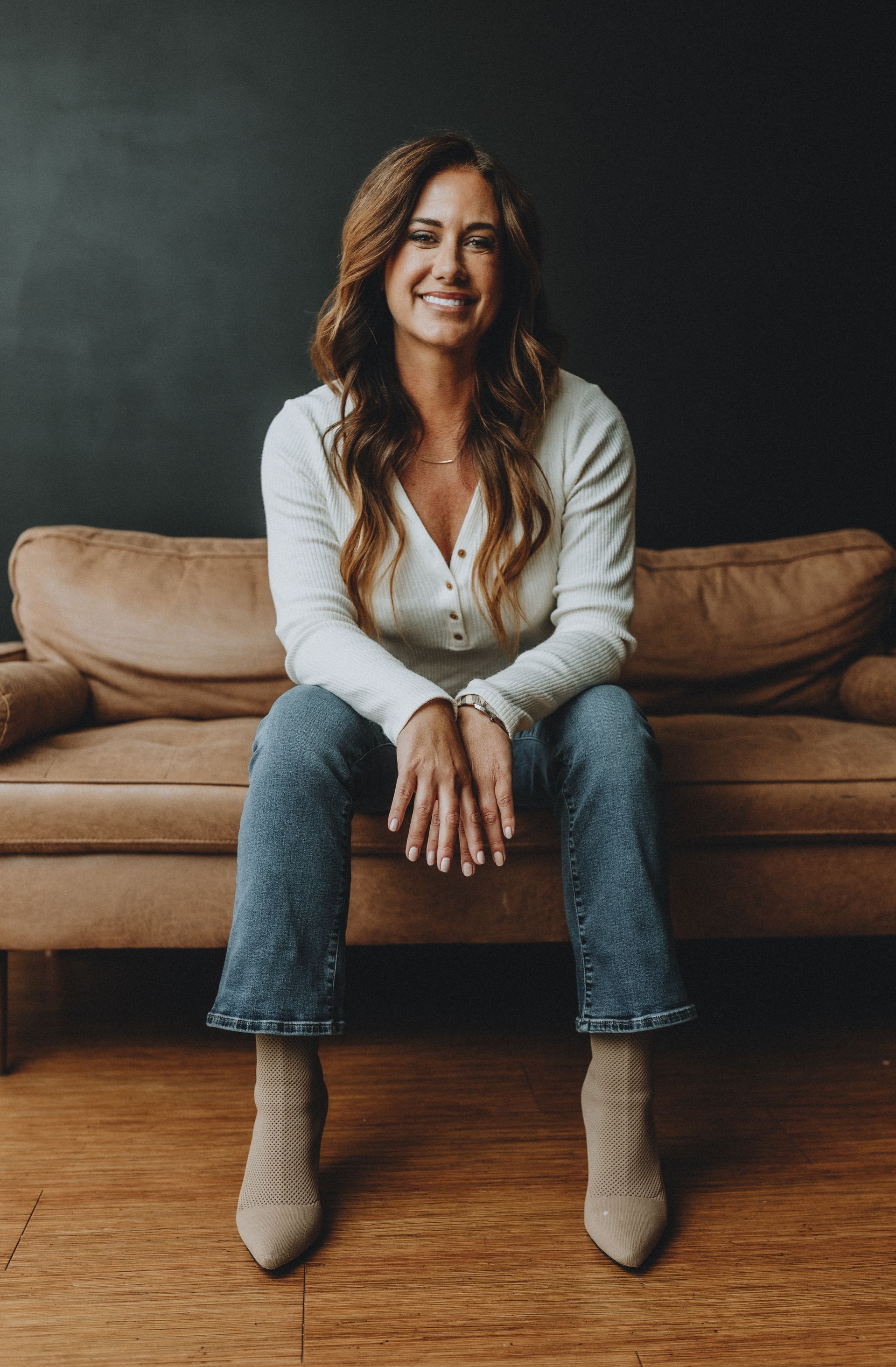 Woman with long brown hair smiles while sitting on a brown couch wearing a white sweater, blue jeans, and tan boots.