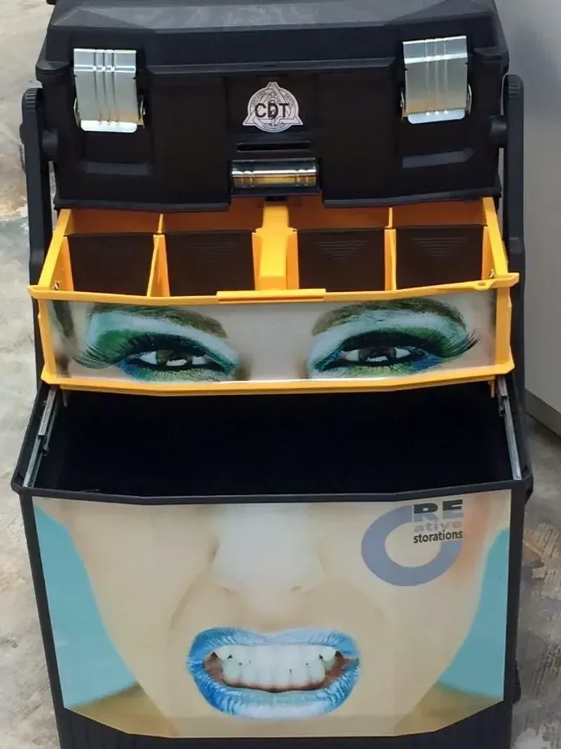 Black toolbox with woman's face on it; open to show drawers and compartments.