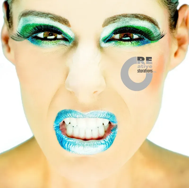 Woman with bold blue and green makeup, white teeth, and a fierce expression.