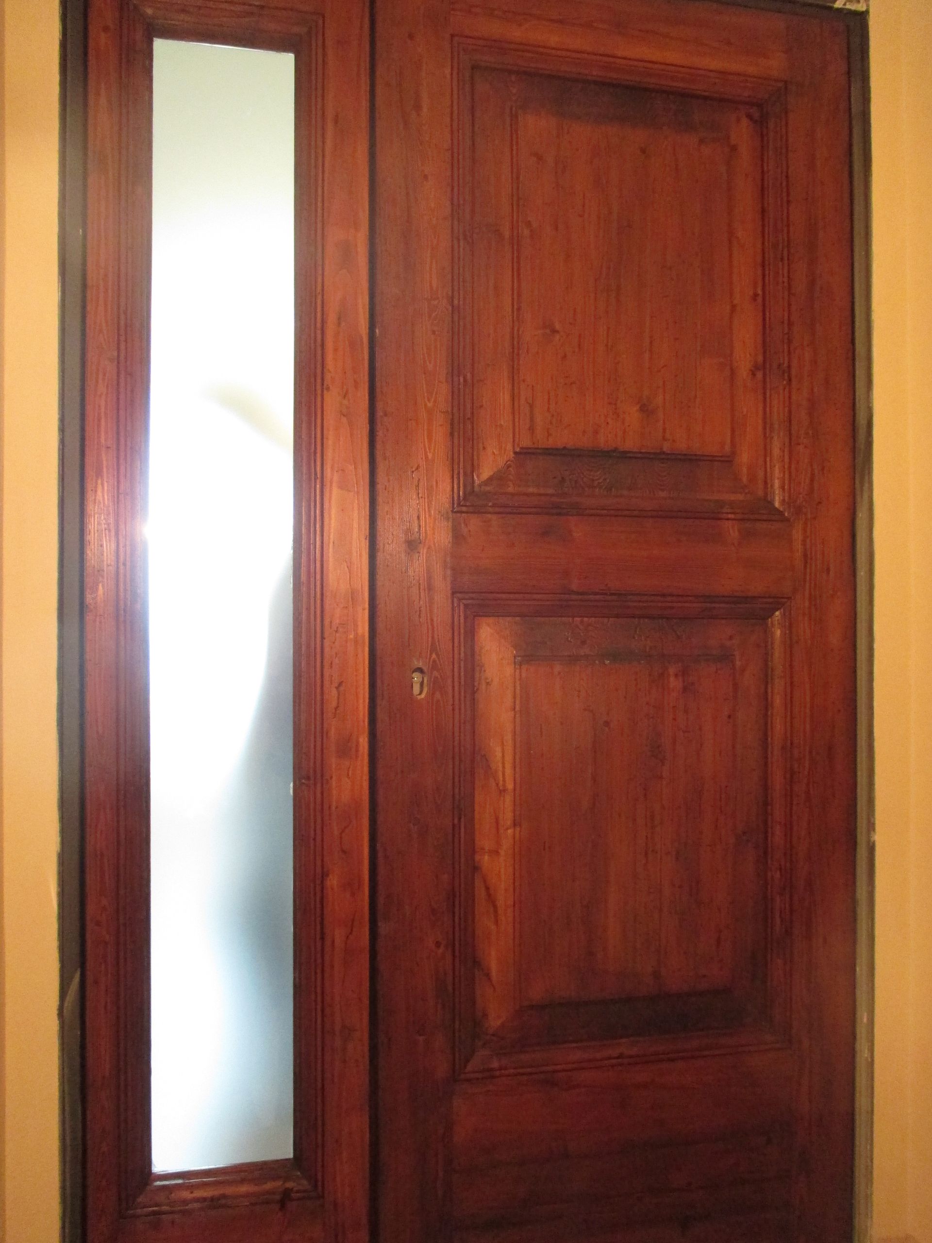 Una porta in legno scuro con due pannelli rettangolari e un pannello laterale verticale in vetro satinato sulla sinistra.