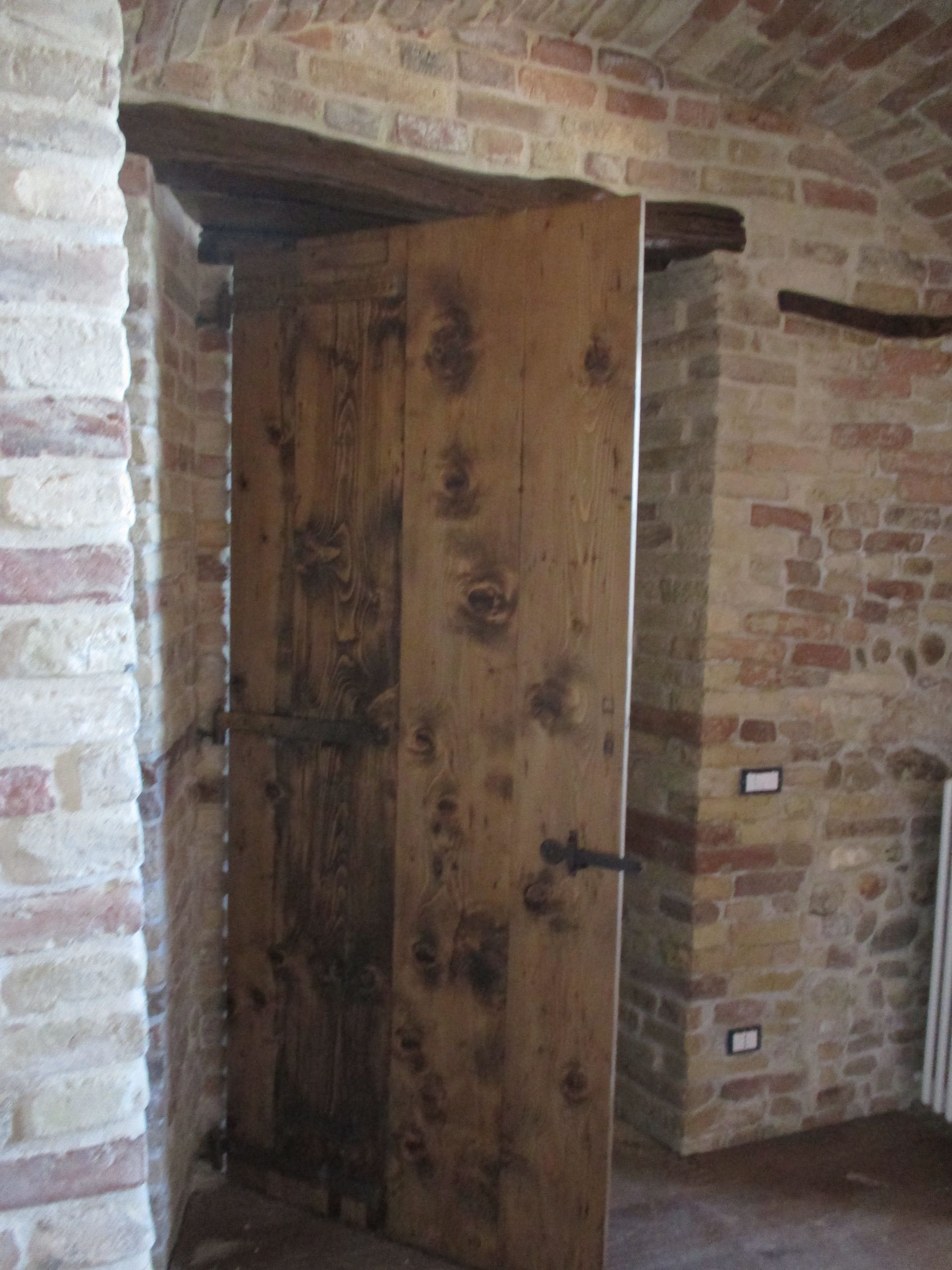 Una porta di legno rustica e nodosa è leggermente socchiusa in un muro di pietra con un arco in mattoni.