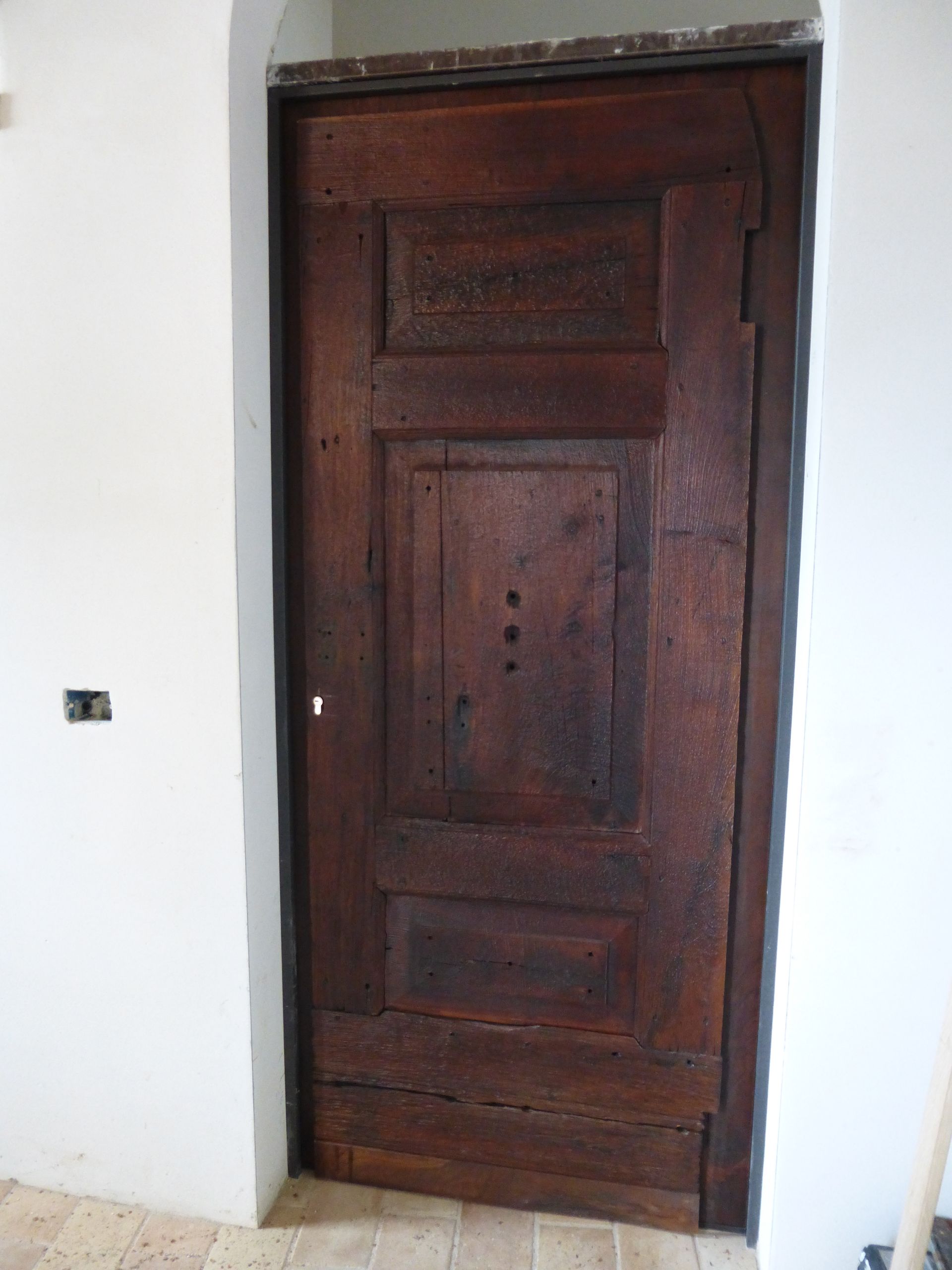 Una porta alta e rettangolare in legno scuro con semplici dettagli rettangolari intagliati, incastonata in una parete ad arco bianca.