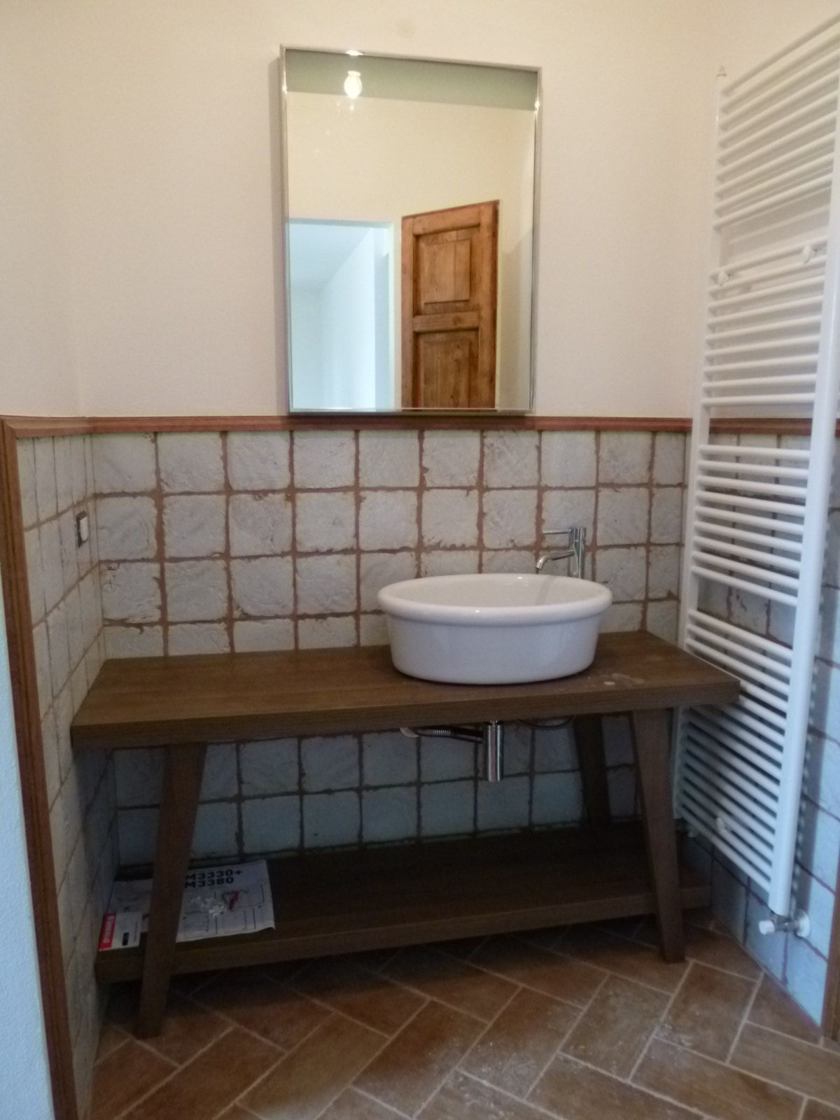 Un lavabo ovale bianco è posizionato su un tavolino consolle in legno rustico, addossato a una parete piastrellata, con specchio e scaldasalviette.