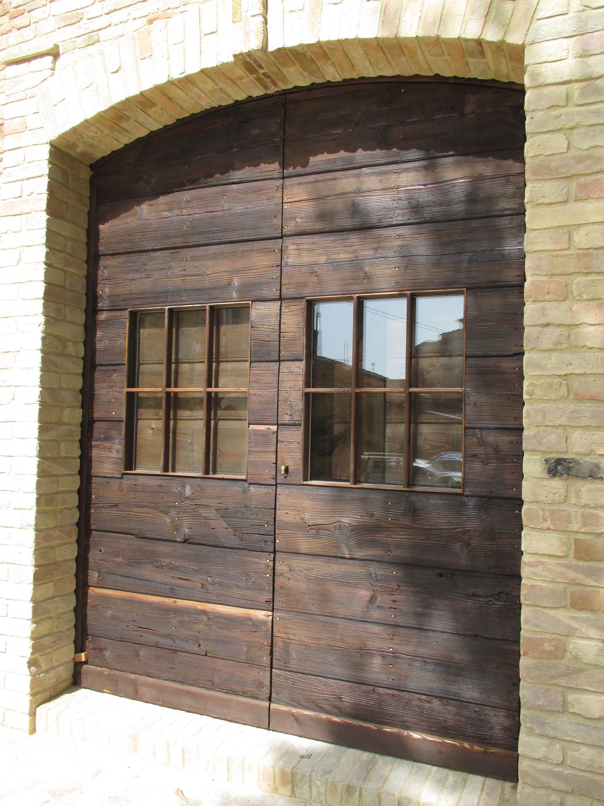Una coppia di doppie porte rustiche in legno scuro con finestre rettangolari, inserite in un ingresso ad arco in mattoni chiari.