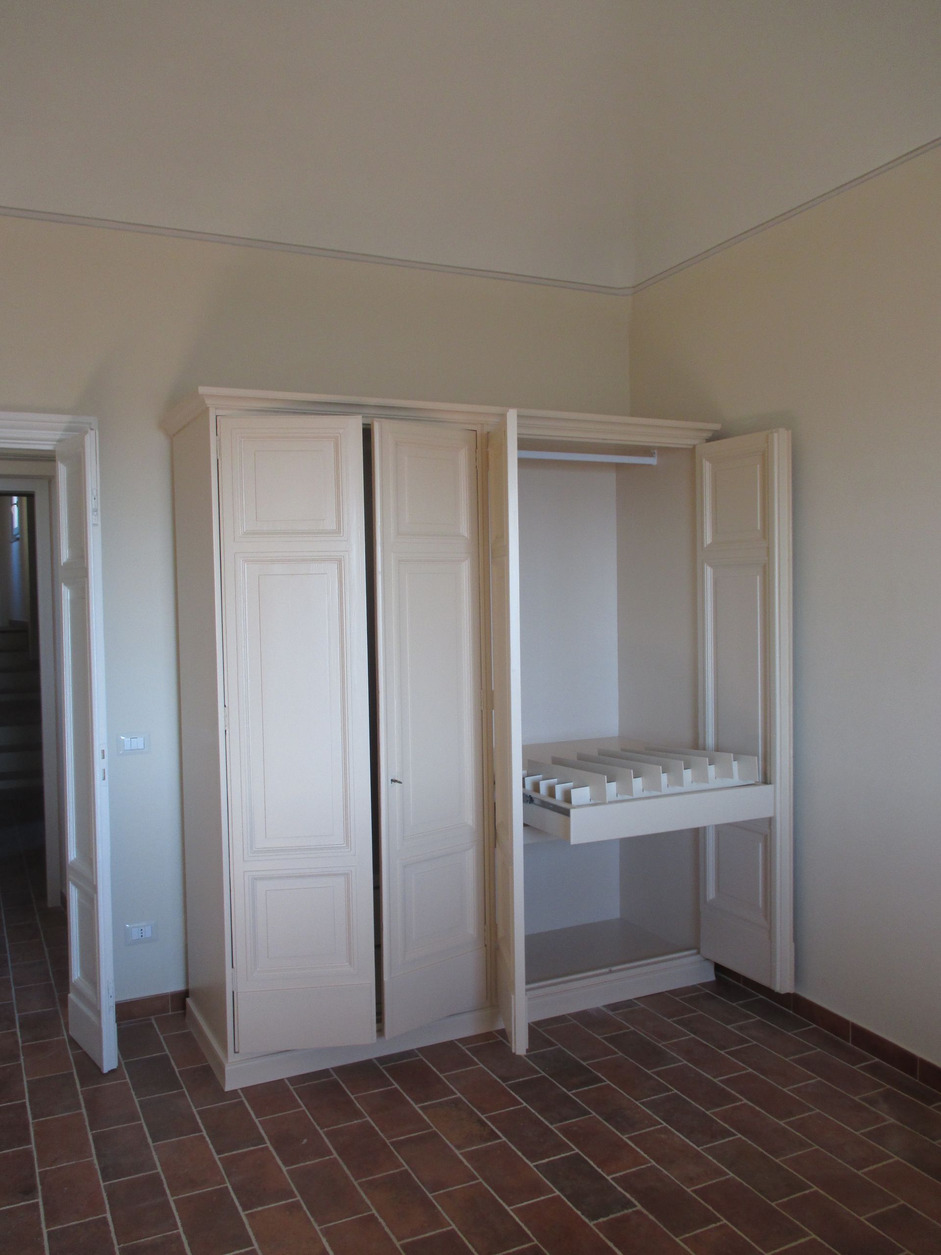 In una stanza con pavimento piastrellato, un armadio in legno chiaro con ante aperte è addossato a una parete.