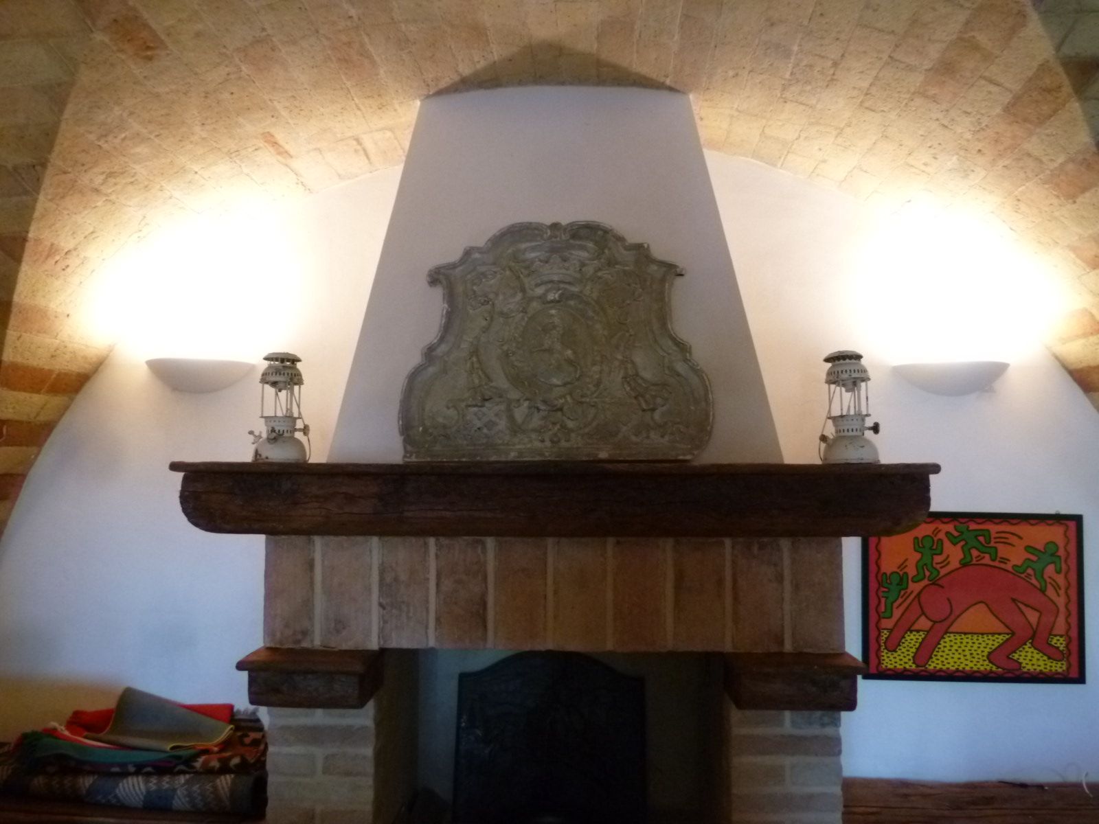 In una stanza con soffitto a volta, si trovano un camino in pietra con un elemento decorativo centrale in metallo, due piccole lanterne e colorati quadri alle pareti.