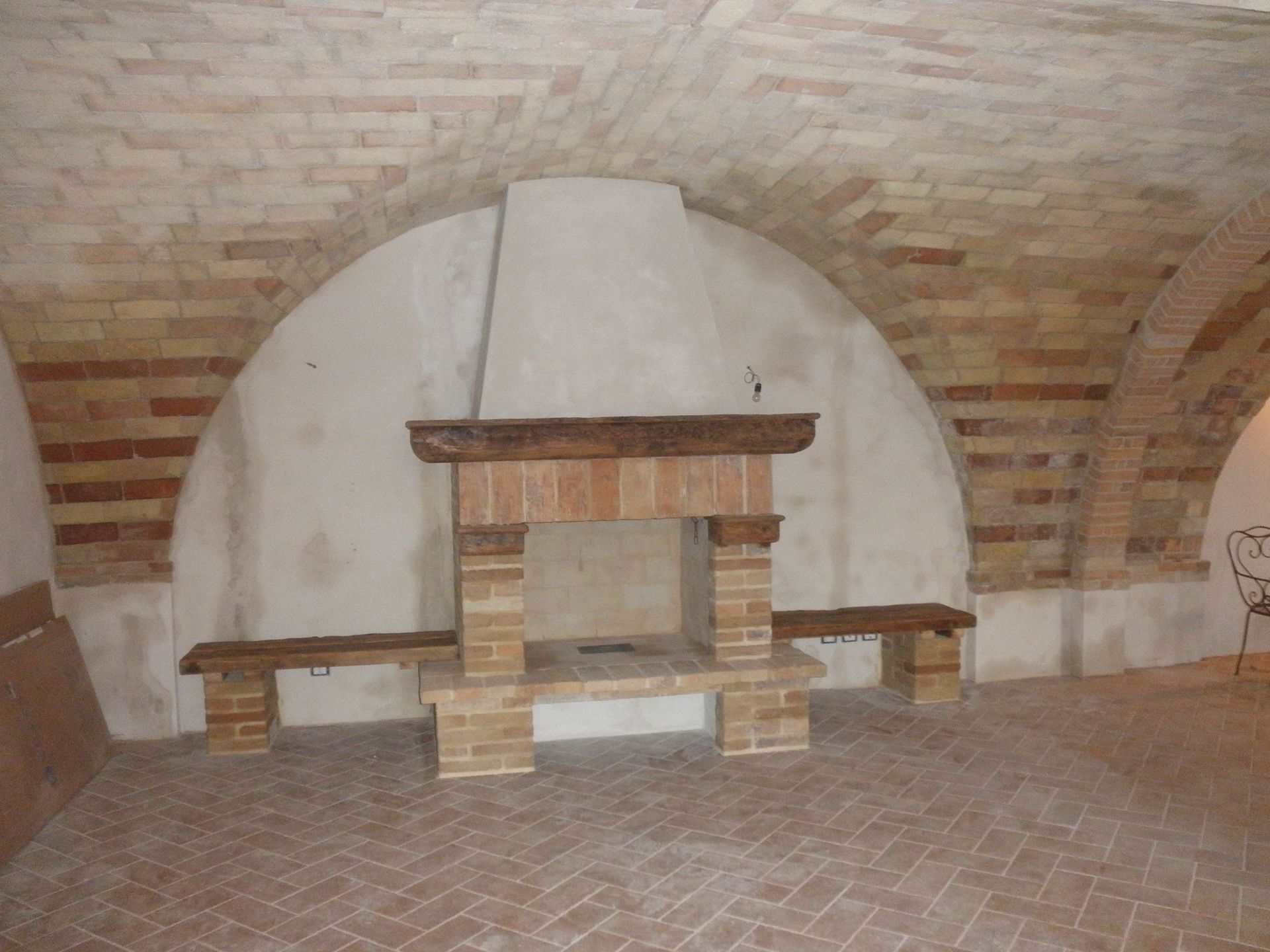 In una stanza rustica con pareti in pietra, sotto un soffitto a volta in mattoni si trova un camino in mattoni con mensola in legno.