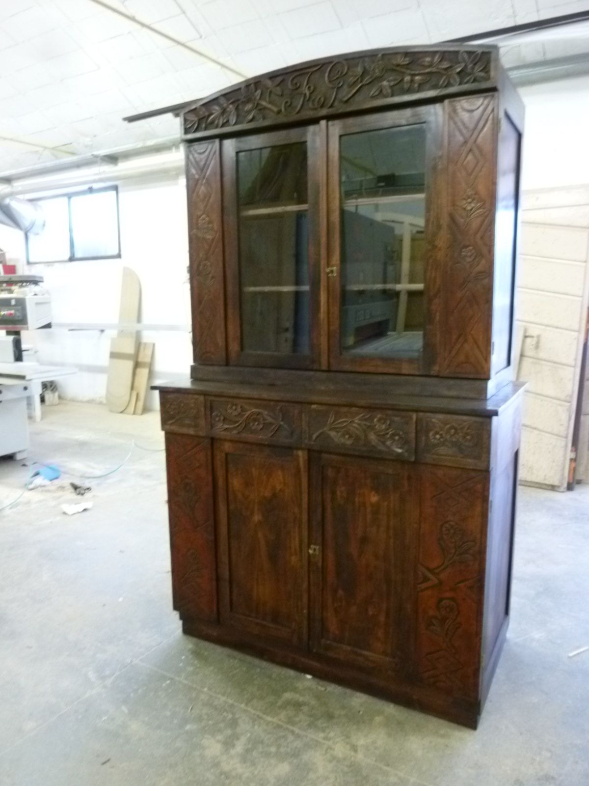 In un'officina, si trova un armadio alto in legno scuro con ante superiori in vetro, intagli elaborati e vani portaoggetti inferiori.