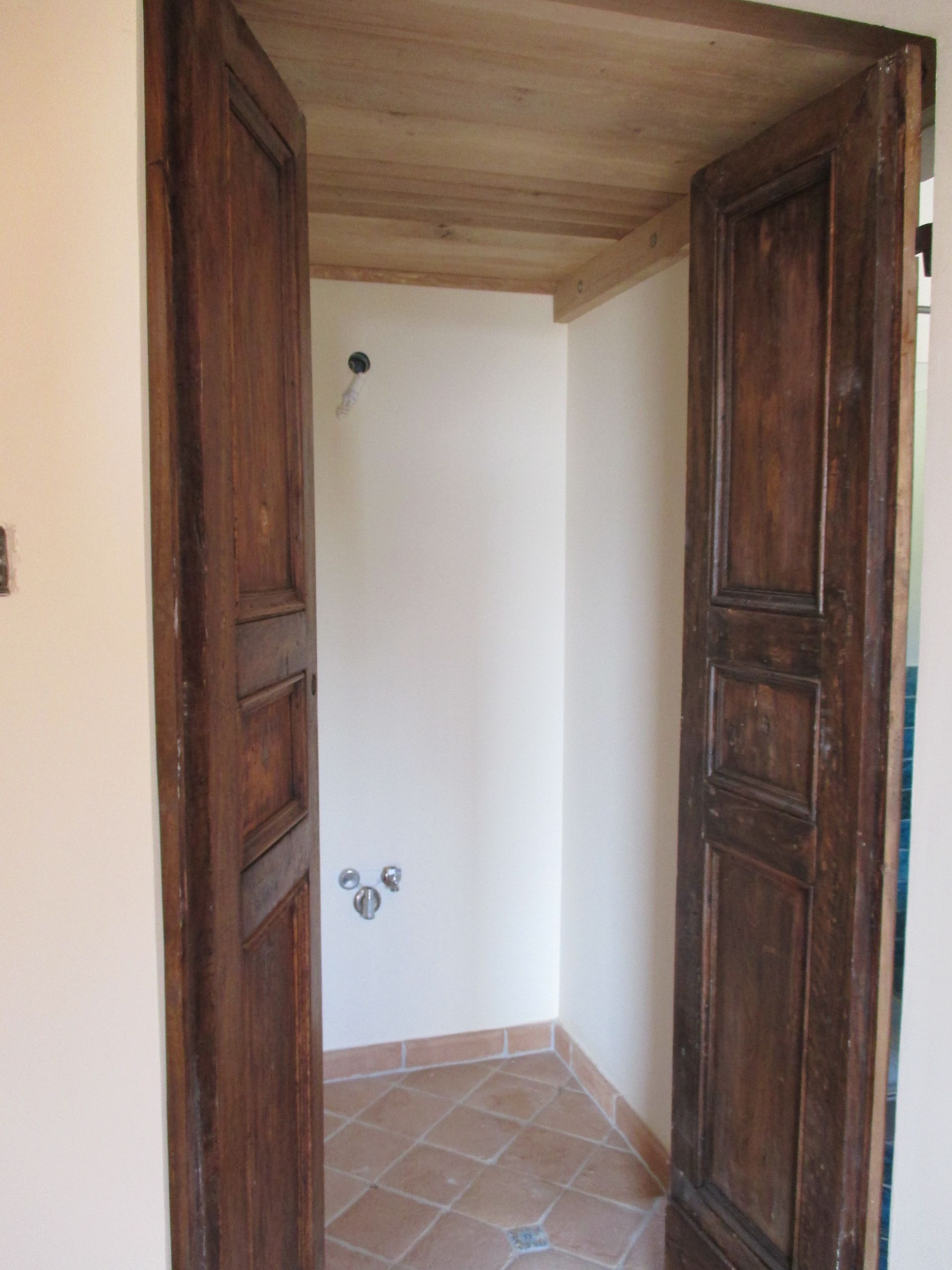 Un ingresso con alte porte a pannelli in legno scuro che si aprono su una piccola stanza piastrellata con sanitari sulla parete di fondo.