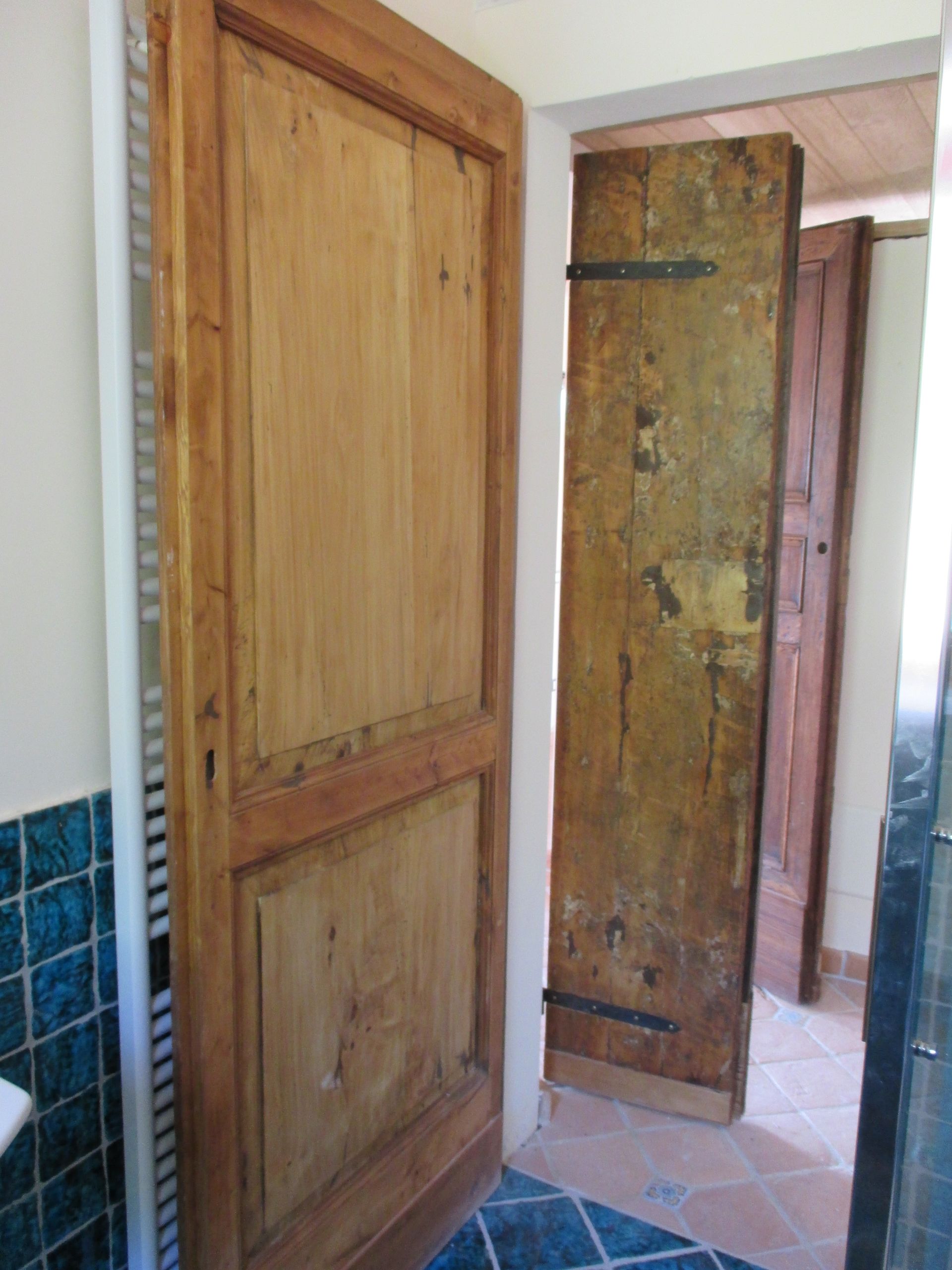 Una porta di legno con due pannelli incassati si trova accanto a un ingresso che lascia intravedere all'interno una porta di legno rustica e invecchiata.