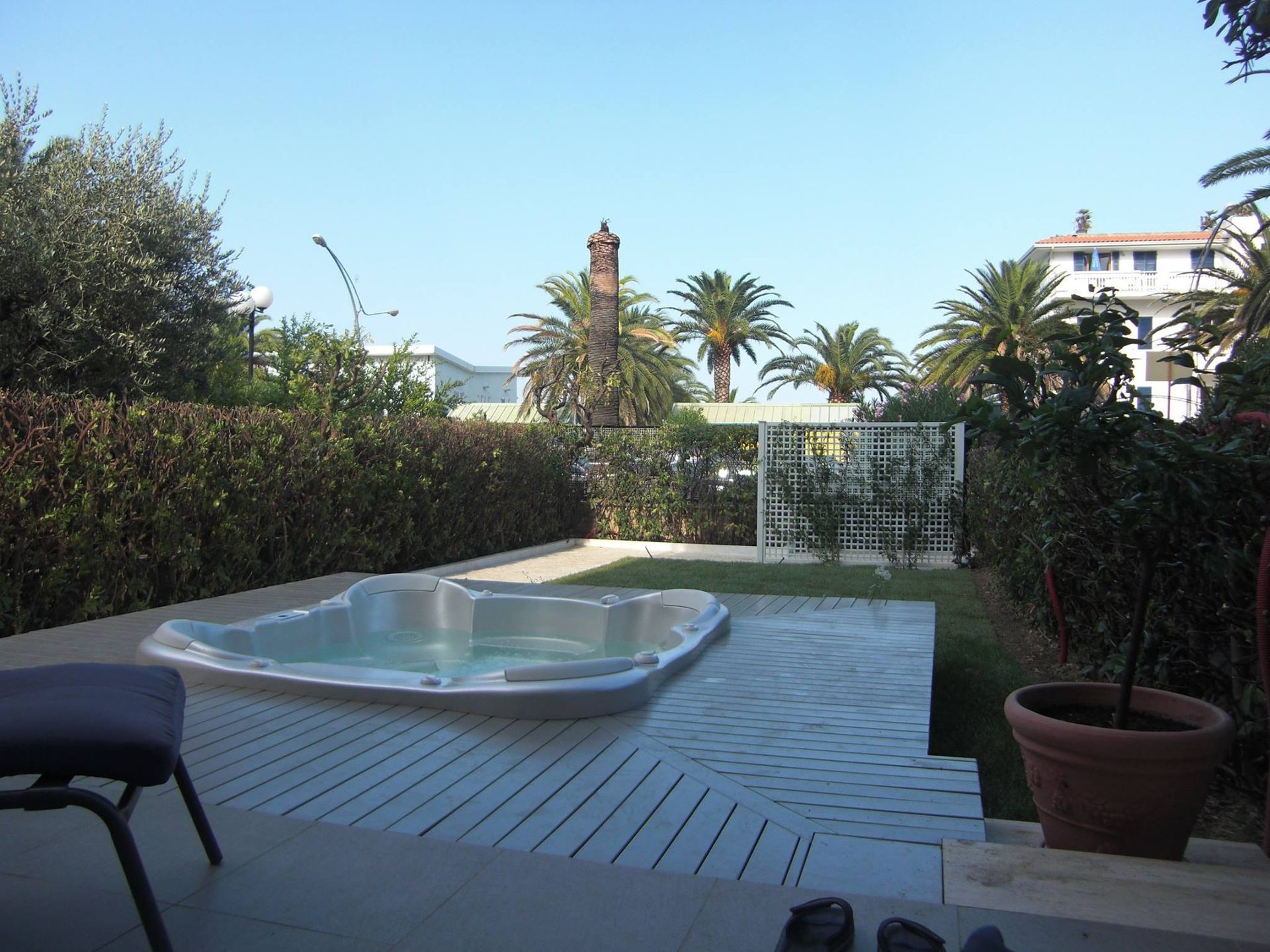 Un patio coperto con una sdraio e una vasca idromassaggio, che si affaccia su un giardino soleggiato con palme e una siepe.