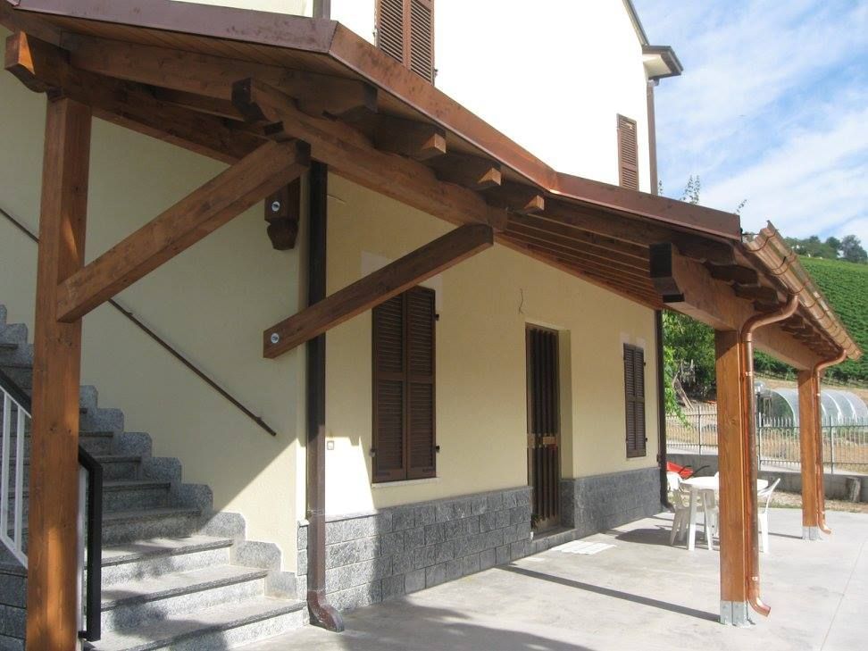 Una casa colonica in pietra a due piani con veranda coperta, scala esterna e piante in vaso sotto un cielo limpido.