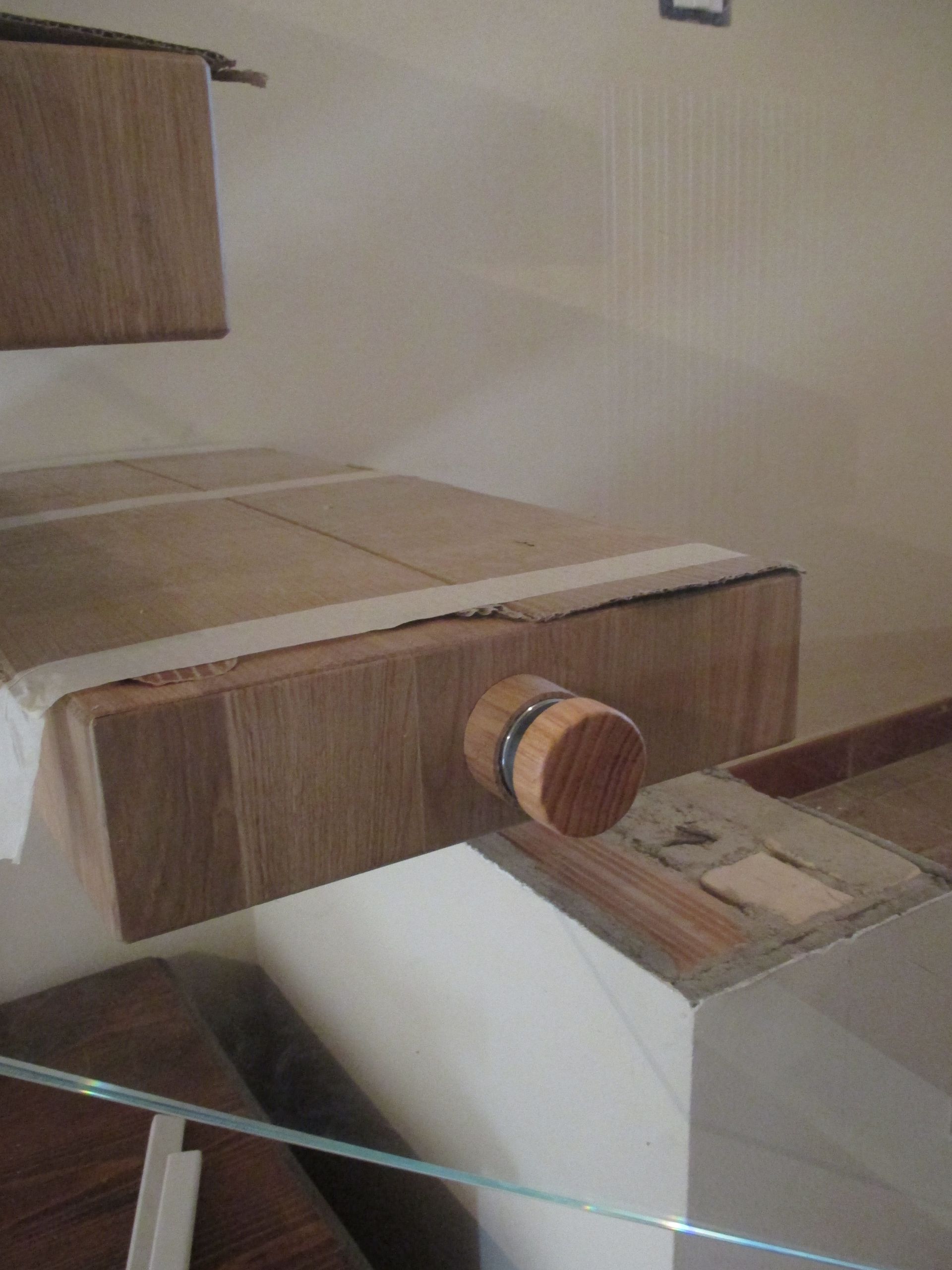 Un elemento di una mensola in legno con pomello circolare, parzialmente installato vicino a una parete con nastro adesivo visibile sui bordi.